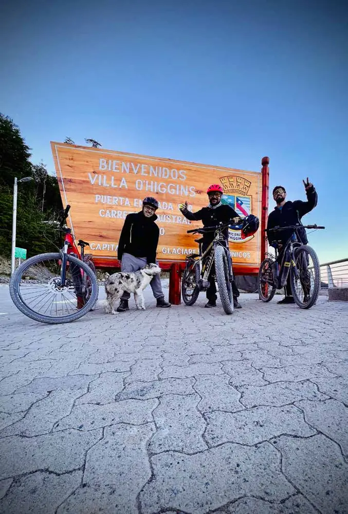 Tres ciclistas con sus bicicletas y un perro junto al cartel de bienvenida de Villa O'Higgins